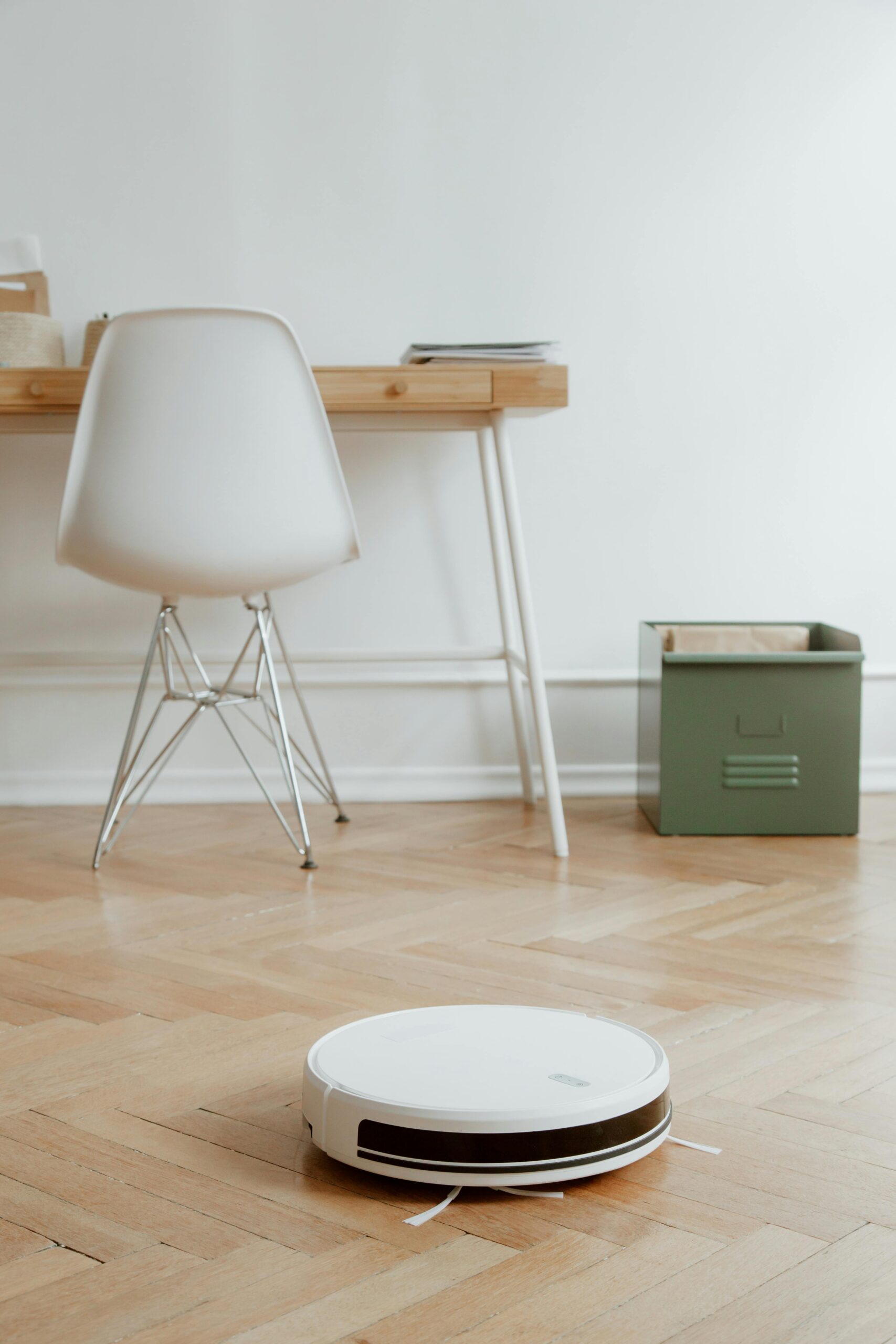A modern minimalist room featuring a robotic vacuum cleaner on a wooden floor beside a desk and chair.